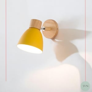 May include: A wall-mounted lamp featuring a bright yellow dome-shaped shade and a light-colored wooden base. The fixture includes a cylindrical wooden connector attached to a circular wooden wall plate, with a metallic joint for adjustable positioning.