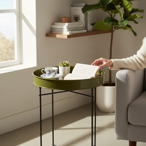 May include: A round, olive green side table with black legs. A teacup and saucer with blue floral designs, a small potted succulent, and an open book rest on the table. A person's arm rests on a gray armchair.