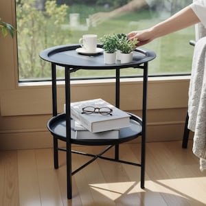 May include: A black metal two-tier round side table with a raised edge on each tray. The top tier holds a white ceramic cup on a saucer and three small potted green plants. The bottom tier holds two stacked white books and a pair of reading glasses.
