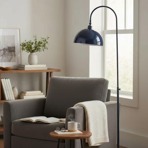 Może przedstawiać: Granatowa lampa podłogowa z łukiem i kloszem w kształcie kopuły oświetla szary fotel. Mały drewniany stolik boczny mieści kubek, książkę i okulary. Kremowy koc jest zarzucony na fotel.