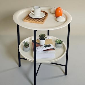 May include: A two-tiered side table with a white tray top and bottom, supported by black metal legs. The top shelf holds a white mug, saucer, and wooden tray. The bottom shelf displays magazines, a remote, and small potted plants.