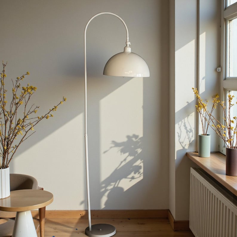 Może przedstawiać: Biała, łukowata lampa podłogowa z zaokrąglonym kloszem. Lampa ma okrągłą podstawę i smukły słup. Pok&oacute;j zawiera st&oacute;ł, krzesło i okno ze światłem słonecznym.
