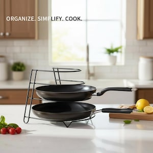 May include: A black metal pan organizer holding two black frying pans. The organizer has three tiers and the text "ORGANIZE. SIMPLIFY. COOK." is at the top. The pans have black handles and are on a white countertop.
