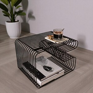 May include: Black metal side table with a two-tiered design. The top shelf holds a cup of coffee and a small square of paper. The lower shelf contains two books. A potted plant is in the background.