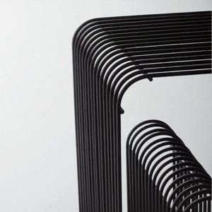 May include: Close-up of a modern, black metal structure with curved and straight lines. The design features a series of parallel, rounded bars that create a geometric pattern against a white background. The minimalist aesthetic suggests a contemporary art piece or furniture design.
