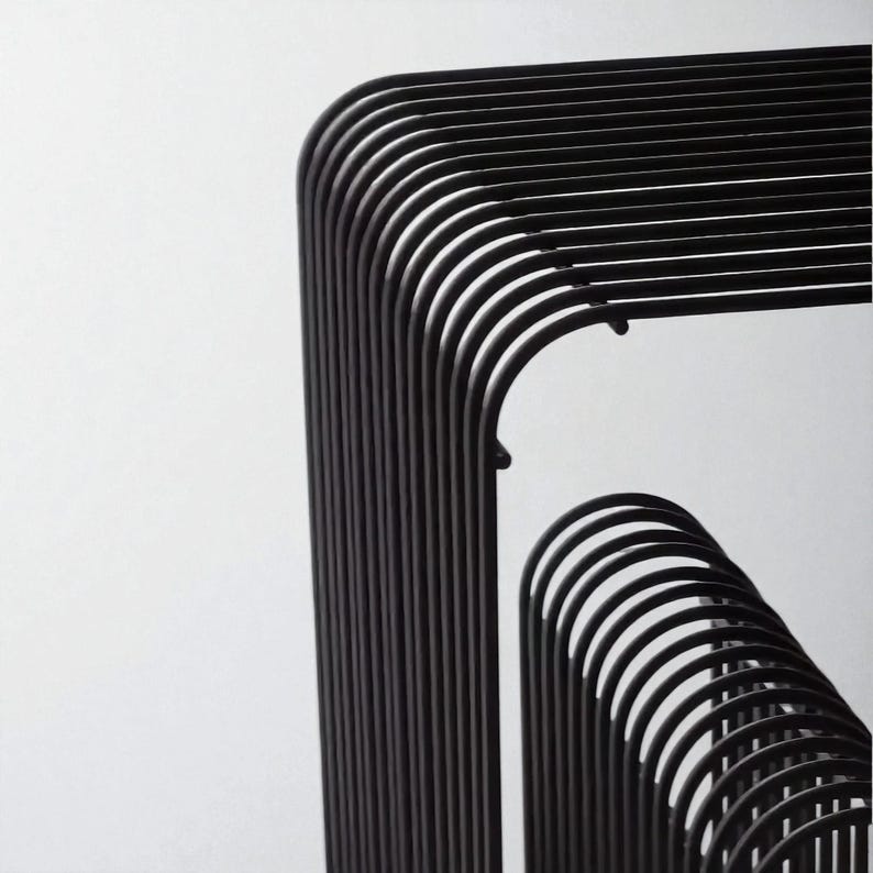 May include: Close-up of a modern, black metal structure with curved and straight lines. The design features a series of parallel, rounded bars that create a geometric pattern against a white background. The minimalist aesthetic suggests a contemporary art piece or furniture design.