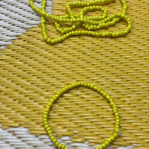 May include: A collection of yellow beaded jewelry, including a long necklace and a bracelet, displayed on a textured yellow and white surface. The beads are small and round, with a consistent color throughout.