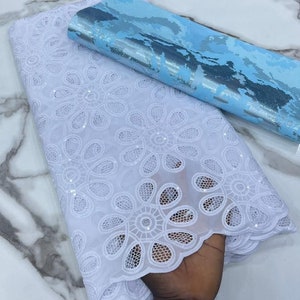 Puede incluir: Tela de encaje blanca con un delicado patrón floral y lentejuelas. La tela tiene un borde festoneado y está superpuesta sobre una tela azul y plateada.