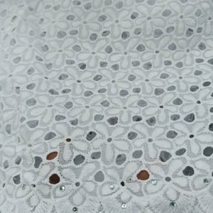 May include: Close-up of white eyelet fabric with a floral pattern. The fabric has a delicate, openwork design with small, round holes and is embellished with tiny, sparkling accents. The texture is soft and intricate, suitable for various sewing projects.