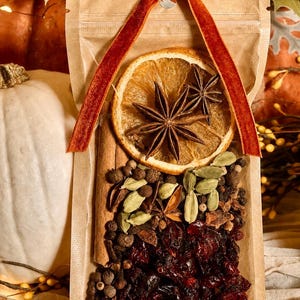 May include: A clear plastic pouch containing a mulling spice blend with a dried orange slice, two star anise, cinnamon sticks, green cardamom pods, whole black peppercorns, and dried cranberries, topped with a rust-colored velvet ribbon.