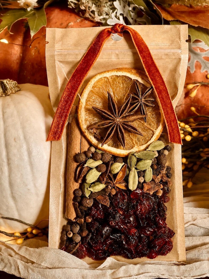 May include: A clear plastic pouch containing a mulling spice blend with a dried orange slice, two star anise, cinnamon sticks, green cardamom pods, whole black peppercorns, and dried cranberries, topped with a rust-colored velvet ribbon.