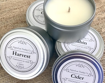 May include: Several silver-colored, round tin candles with white labels are arranged on a textured surface. One candle is open, revealing a cream-colored wax and a wick. The labels read "Harvest," "Cabin," and "Cider," with the text "Hand-poured soy candle" and "5 ounces."