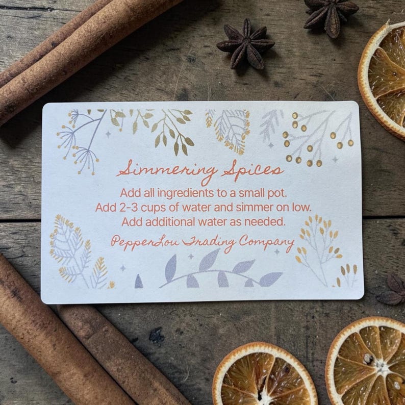 May include: A rectangular white card with rounded corners featuring orange text that reads Simmering Spices, instructions to add ingredients to a small pot with 2-3 cups of water, and the brand name PepperLou Trading Company, decorated with botanical illustrations.