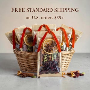 May include: A woven wicker basket filled with several clear pouches containing mulling spices, including dried orange slices, star anise, cinnamon sticks, green cardamom pods, and dark red dried cranberries, each tied with a bright orange velvet ribbon.