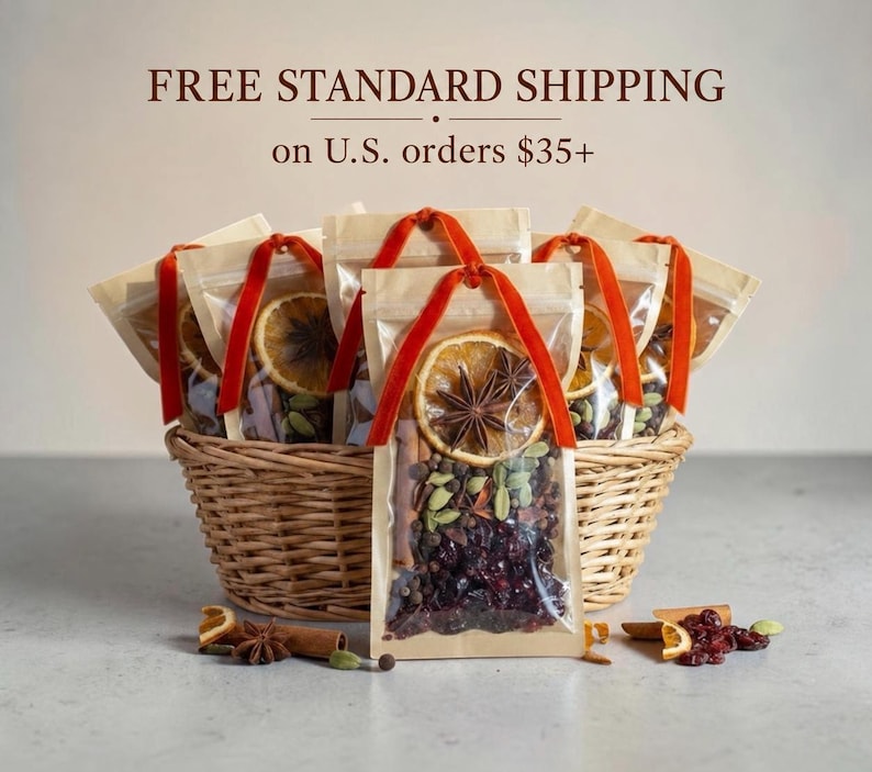 May include: A woven wicker basket filled with several clear pouches containing mulling spices, including dried orange slices, star anise, cinnamon sticks, green cardamom pods, and dark red dried cranberries, each tied with a bright orange velvet ribbon.