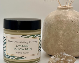 May include: A glass jar of Lavender Tallow Balm with a black lid, labeled "Pepperlou Trading Company." The jar is next to a beige vase with dried flowers and a small, light-colored stone. The balm weighs 4 ounces.