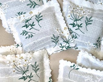 May include: A collection of rectangular fabric sachets featuring a white base with green botanical prints of wildflowers and foliage. Each sachet has scalloped edges, intricate lace trim detailing, and a textured surface with small white floral accents.