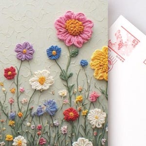 May include: A postcard with a textured design of crocheted flowers in various colours. The flowers include pink, purple, blue, red, yellow, and white blooms with green stems. A blank postcard is next to the floral card.