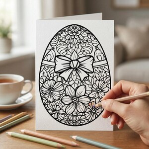 K&ouml;nnte beinhalten: Eine wei&szlig;e Osterkarte mit einem schwarzen floralen Eierdesign und einer Schleife. Eine Hand bemalt das Ei mit einem Buntstift. Buntstifte und eine Tasse Tee stehen auf dem Holztisch.