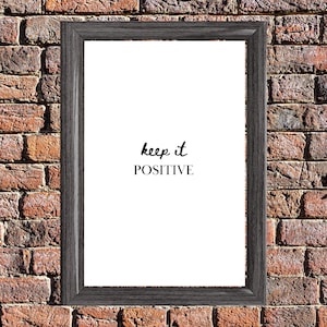 May include: A framed print with the words "keep it POSITIVE" in black script on a white background. The frame is dark gray and has a wood-grain texture. The background is a brick wall.