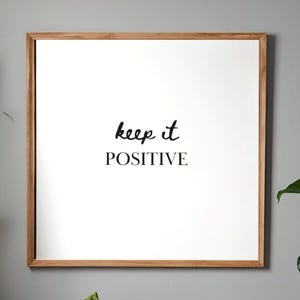 May include: Square framed art print with the words "keep it POSITIVE" in black script on a white background. The frame is made of light brown wood. The artwork is displayed against a gray wall.