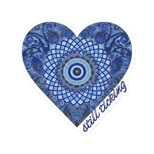 May include: Heart-shaped sticker with a blue mandala design and the words "still ticking" in a handwritten font. The mandala features intricate patterns in shades of blue and a central eye-like design.