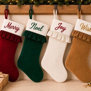 May include: Four velvet Christmas stockings in deep red, dark green, cream, and tan. Each stocking features a white cuff with embroidered text reading Merry, Noel, Joy, and Wish, respectively, and a matching ruffled trim below the cuff.