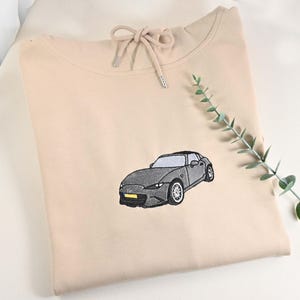 May include: A beige hoodie with a drawstring neckline and a detailed embroidered image of a gray sports car. The car has black tires and a yellow license plate. A sprig of greenery is placed on the right side.