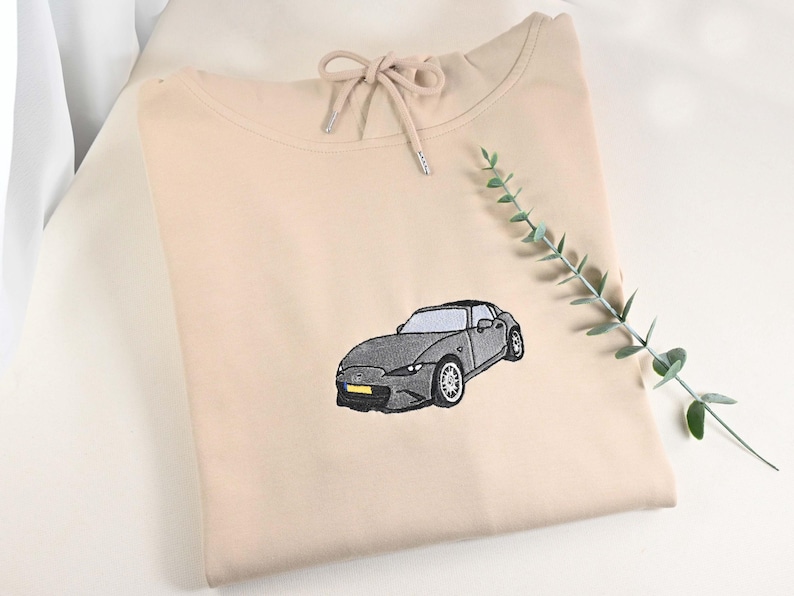 May include: A beige hoodie with a drawstring neckline and a detailed embroidered image of a gray sports car. The car has black tires and a yellow license plate. A sprig of greenery is placed on the right side.