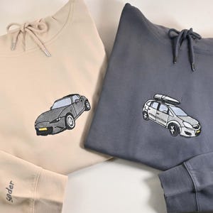 Op de afbeelding: Twee hoodies, één beige en één antracietgrijs. Elk heeft een geborduurd autodesign. De beige hoodie heeft een sportwagen en de grijze heeft een auto met een dakrek. De manchet van de grijze hoodie leest "2025".