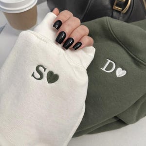 May include: Close-up of two sweatshirts, one white and one olive green. The white sweatshirt sleeve has an embroidered "S" and a heart. The green sweatshirt has a "D" and a heart. The image also shows a hand with black nail polish.