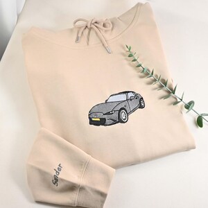 May include: Beige hoodie with a drawstring neckline and a gray car embroidered on the front. The left sleeve has the name "Sander" embroidered in a matching color. A sprig of greenery is placed on the right side of the hoodie.