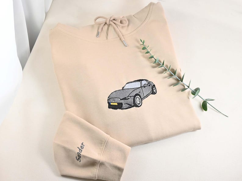 May include: Beige hoodie with a drawstring neckline and a gray car embroidered on the front. The left sleeve has the name "Sander" embroidered in a matching color. A sprig of greenery is placed on the right side of the hoodie.