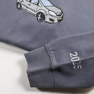 May include: A gray sweatshirt with an embroidered car design. The sleeve cuff displays the year "2025" in white stitching. The sweatshirt is a muted gray color, with a light beige fabric visible in the background.