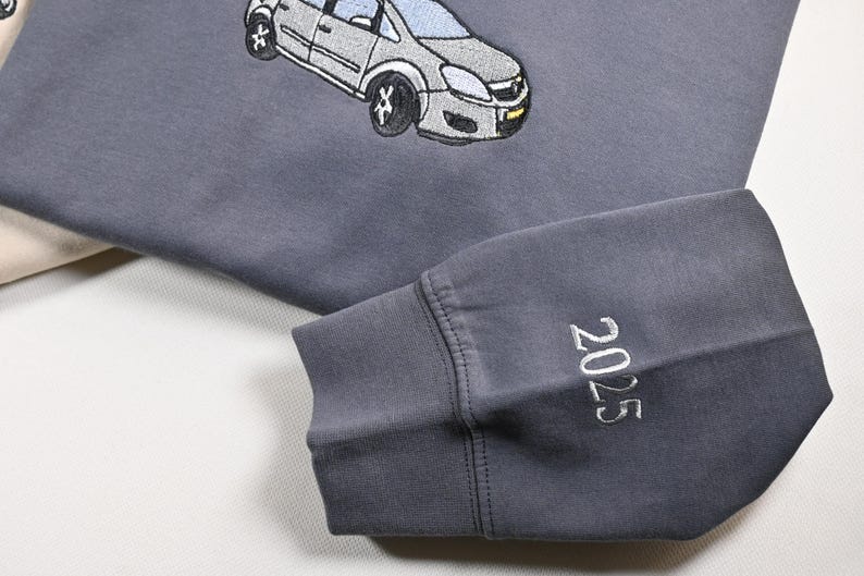 May include: A gray sweatshirt with an embroidered car design. The sleeve cuff displays the year "2025" in white stitching. The sweatshirt is a muted gray color, with a light beige fabric visible in the background.