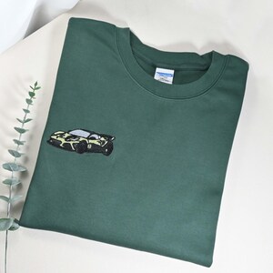 May include: Dark green sweatshirt with a small embroidered car design on the front. The car is black, white, and yellow. The sweatshirt is folded on a white surface with a sprig of greenery.