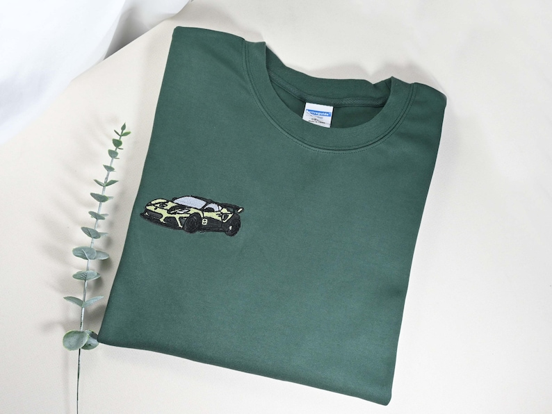 May include: Dark green sweatshirt with a small embroidered car design on the front. The car is black, white, and yellow. The sweatshirt is folded on a white surface with a sprig of greenery.