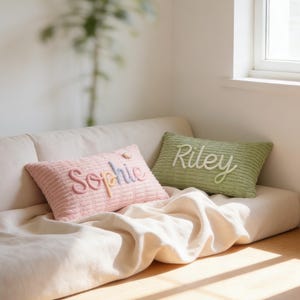 May include: Two rectangular corduroy cushions on a beige sofa. One is light pink with the name "Sophie" embroidered in pastel colours. The other is light green with the name "Riley" embroidered in white. A cream-coloured blanket is draped over the sofa.