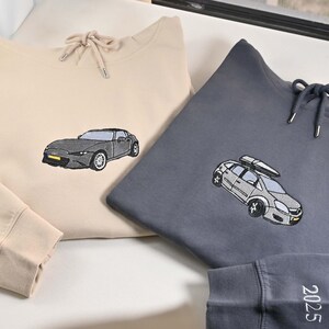 May include: Two hooded sweatshirts, one beige and one gray, with embroidered car designs. The beige sweatshirt has a car graphic, and the gray one features a car with a roof rack. The beige sleeve has the word "sinder" embroidered on it, and the gray sleeve has the number "2025".