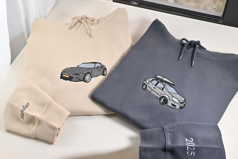 May include: Two hooded sweatshirts, one beige and one gray, with embroidered car designs. The beige sweatshirt has a car graphic, and the gray one features a car with a roof rack. The beige sleeve has the word "sinder" embroidered on it, and the gray sleeve has the number "2025".