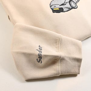 May include: Beige sweatshirt with an embroidered car graphic. The sleeve cuff features the name "Sander" in a matching thread color. The design is simple and clean, suitable for casual wear.