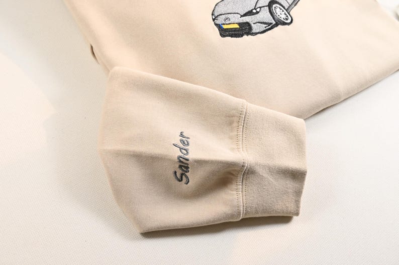 May include: Beige sweatshirt with an embroidered car graphic. The sleeve cuff features the name "Sander" in a matching thread color. The design is simple and clean, suitable for casual wear.