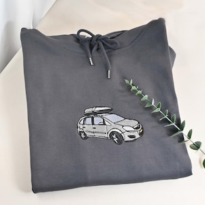 May include: A charcoal gray hoodie with a drawstring hood. The front features an embroidered design of a silver car with a roof rack. A sprig of greenery is placed to the right of the hoodie.