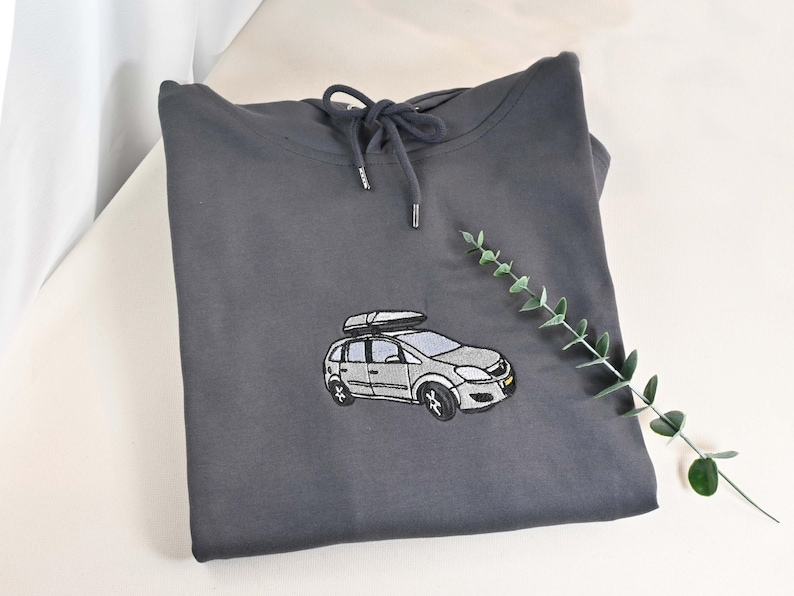 May include: A charcoal gray hoodie with a drawstring hood. The front features an embroidered design of a silver car with a roof rack. A sprig of greenery is placed to the right of the hoodie.