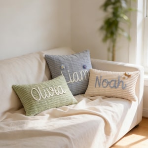 Könnte beinhalten: Drei Cord-Zierkissen mit gestickten Namen. Die Kissen sind in Grün-, Blau- und Cremetönen gehalten. Die Namen "Olivia", "Liam" und "Noah" sind in Schreibschrift dargestellt. Dekorative Blumen und ein Löwe sind ebenfalls vorhanden.