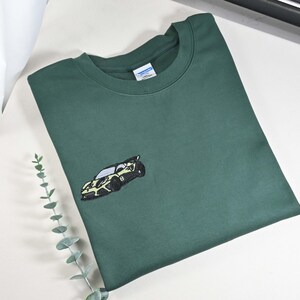 May include: A dark green sweatshirt with a crew neck and an embroidered car design on the front. The car is black and yellow. The sweatshirt is made of a soft material.