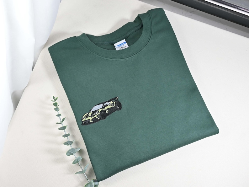 May include: A dark green sweatshirt with a crew neck and an embroidered car design on the front. The car is black and yellow. The sweatshirt is made of a soft material.