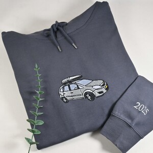 May include: A dark gray hoodie with a drawstring hood and a car graphic embroidered on the front. The car has a roof rack. The sleeve cuff has the embroidered number "2025". A sprig of greenery is in the background.