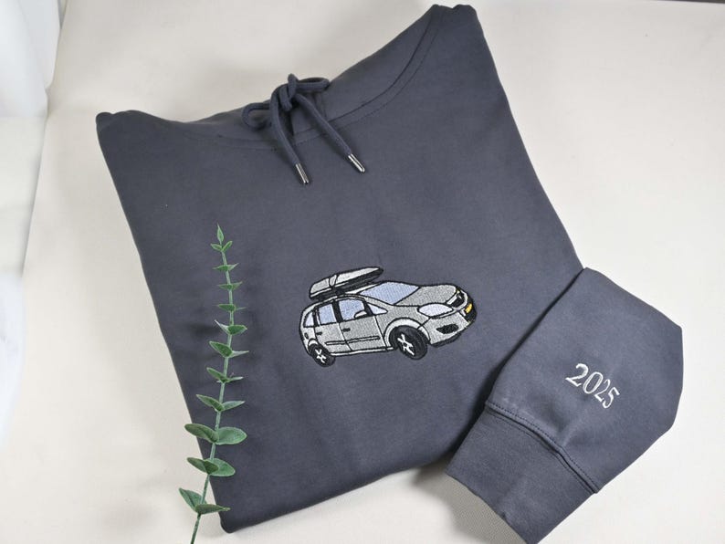 May include: A dark gray hoodie with a drawstring hood and a car graphic embroidered on the front. The car has a roof rack. The sleeve cuff has the embroidered number "2025". A sprig of greenery is in the background.