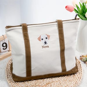 May include: Cream-colored tote bag with brown accents, featuring an embroidered dog face and the name "Nova." The bag has brown handles and a brown base. A wooden calendar and flowers are in the background.